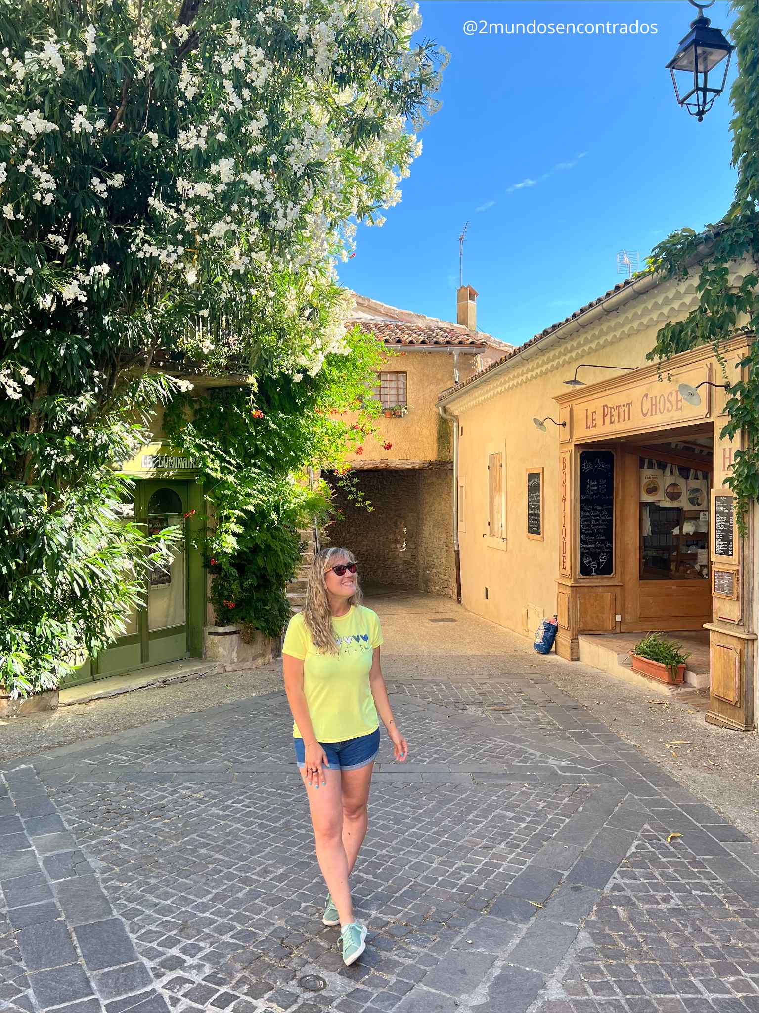 Eva sonriente camina por una calle empedrada y estrecha del pintoresco pueblo de Venasque, en la región de Provenza. A la derecha se ve una tienda tradicional con fachada de madera clara llamada "Le Petit Chose", y a la izquierda una gran enredadera con flores blancas cubre parte de la fachada de los edificios antiguos.