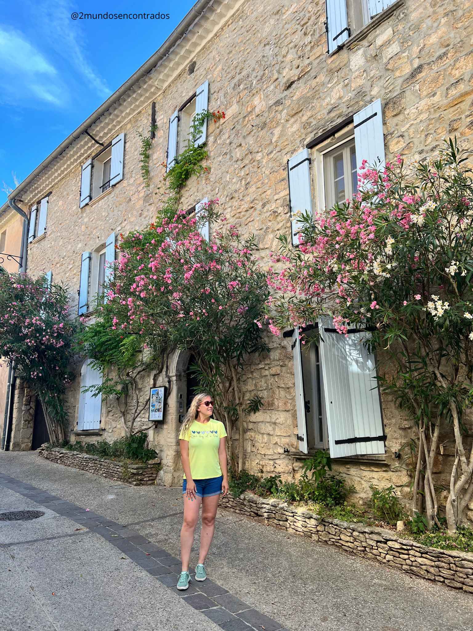 Eva parada junto a un gran edificio tradicional de piedra rústica en Venasque, Francia. La fachada cuenta con contraventanas de madera en un tono azul claro pastel, y está decorada con grandes arbustos de adelfas florecidas con flores rosas y blancas que suben por las paredes coloniales.
