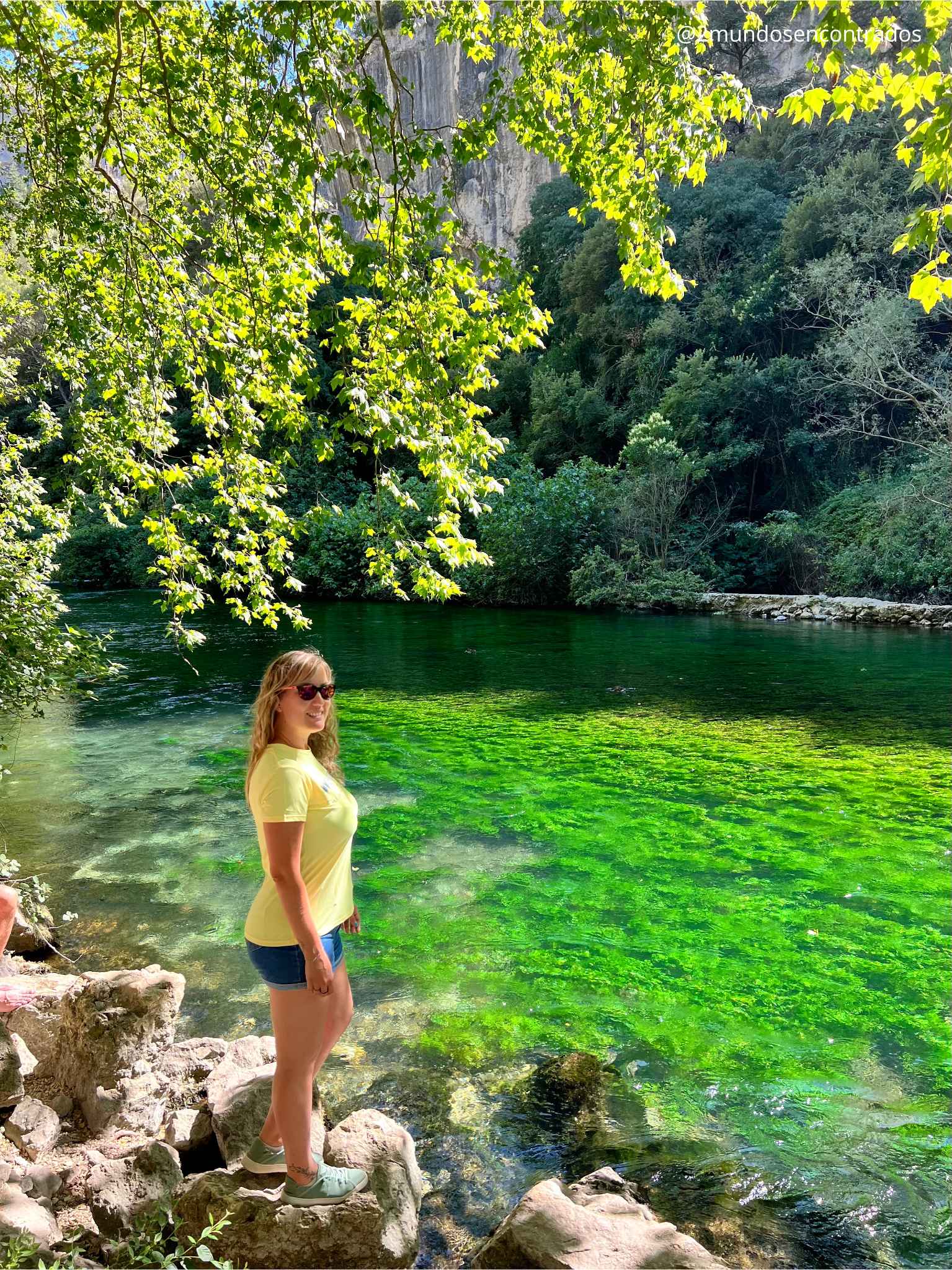 Eva está parada sobre unas rocas junto a la orilla del río Sorgue en Fontaine-de-Vaucluse, Francia. El agua es cristalina con un tono verde vibrante debido a la vegetación subacuática, rodeada de árboles verdes y un acantilado de piedra caliza al fondo.