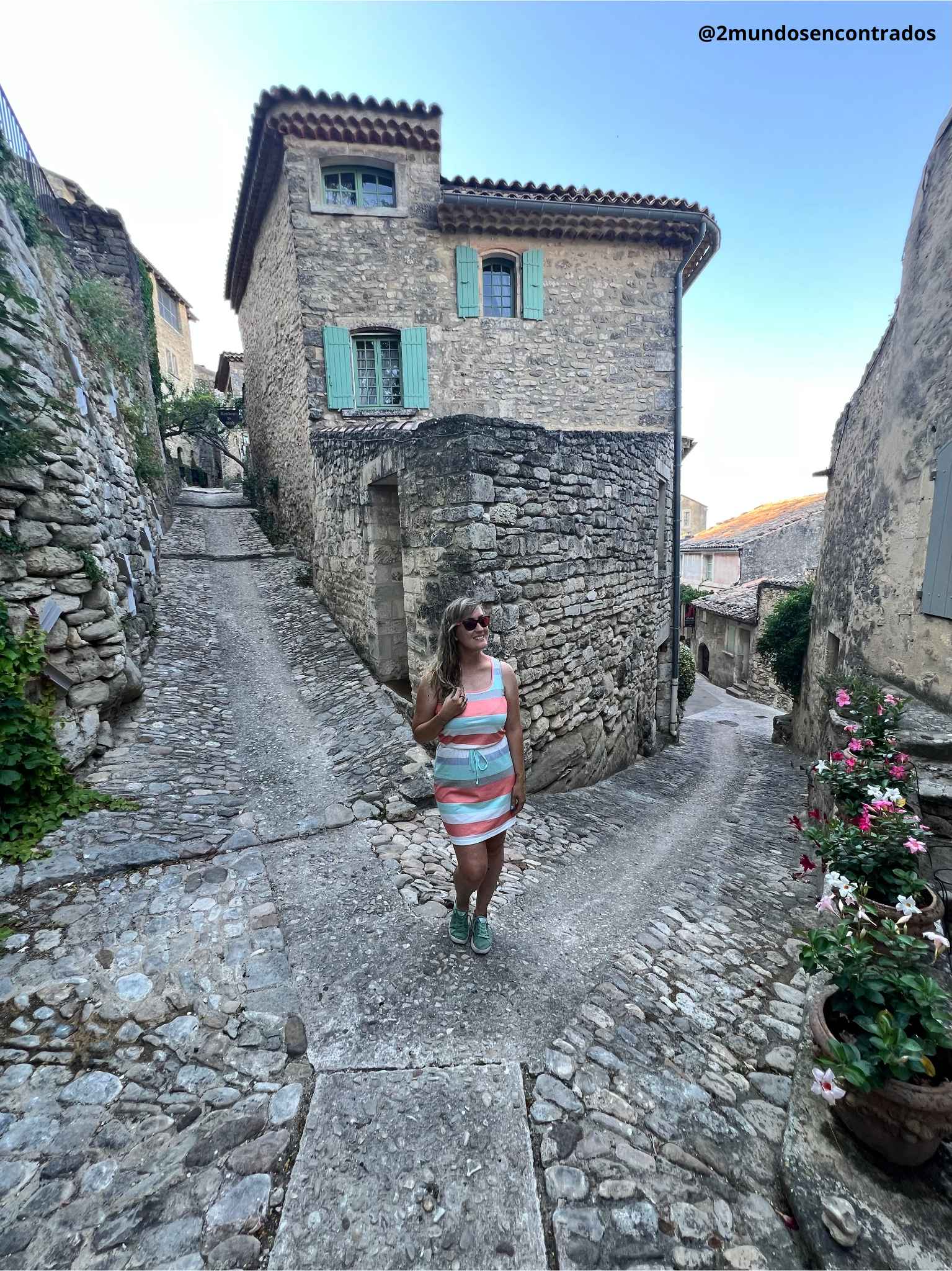 Eva en un cruce de calles empedradas junto a una casa de piedra con contraventanas turquesas en Lacoste.