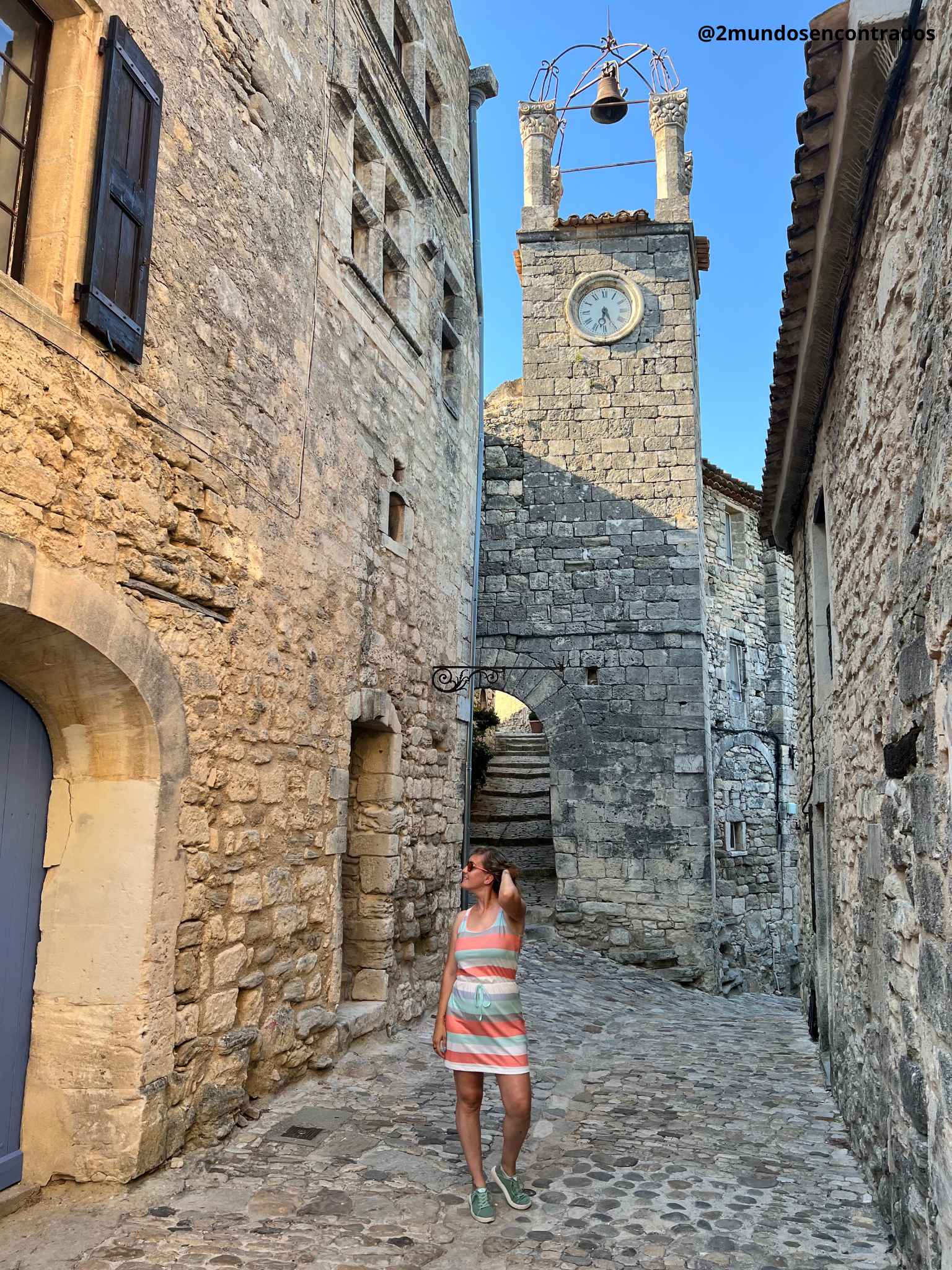 Eva en un callejón empedrado frente a la emblemática Torre del Reloj (Beffroi) de Lacoste, una estructura de piedra antigua con un arco y un campanario de forja bajo un cielo azul despejado.