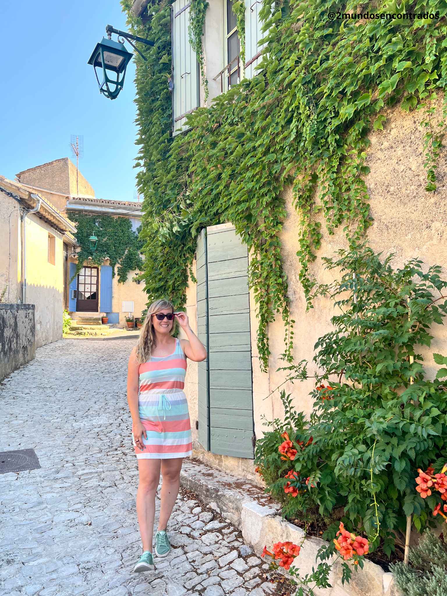 Eva posa junto a una fachada de piedra tradicional en Ménerbes, Provence, caracterizada por una frondosa enredadera verde que cubre la pared, contraventanas de madera de color verde claro y flores de tonos anaranjados en primer plano.