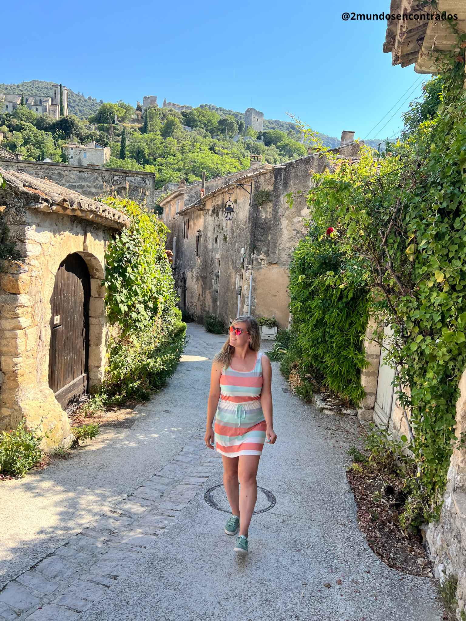 Eva camina por una pintoresca calle empedrada en el pueblo medieval de Oppède-le-Vieux, rodeada de casas de piedra antiguas cubiertas de vegetación y con las ruinas del castillo en lo alto de la colina al fondo.