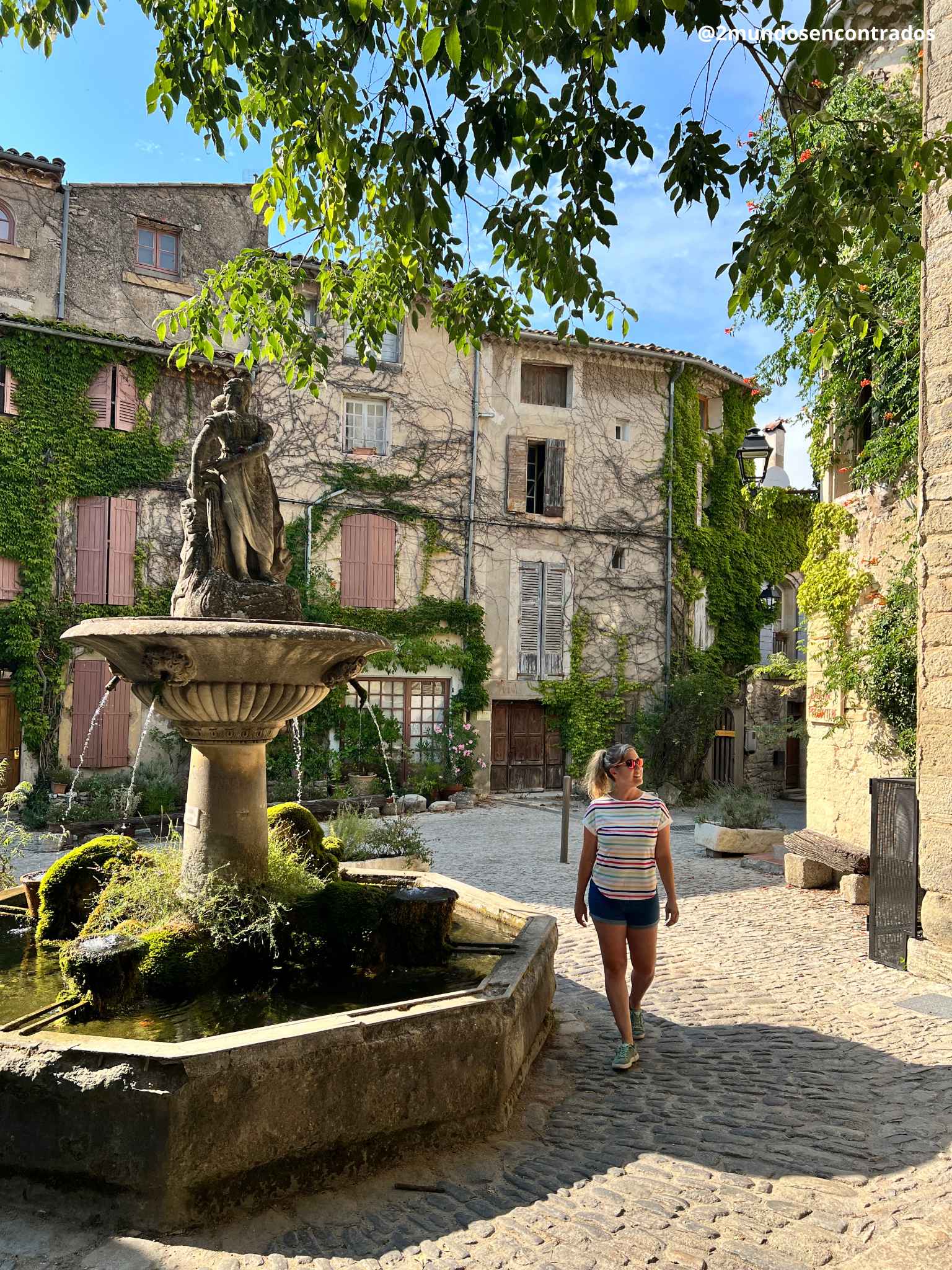 Eva paseando junto a la emblemática fuente de piedra rodeada de fachadas con hiedra en Saignon, Provenza.