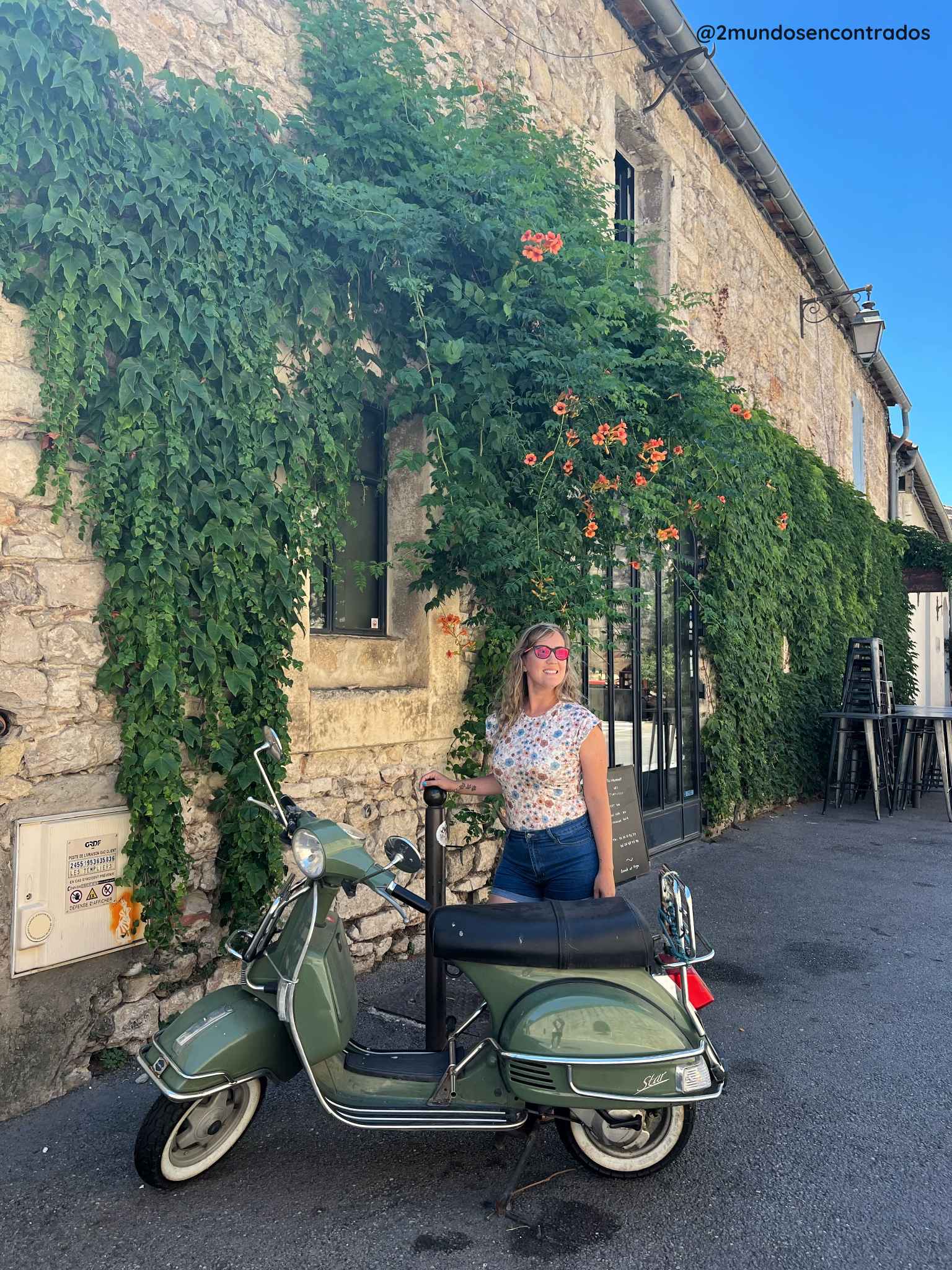 Eva posando junto a una moto clásica de color verde en una calle empedrada frente a una fachada cubierta de vegetación y flores. 