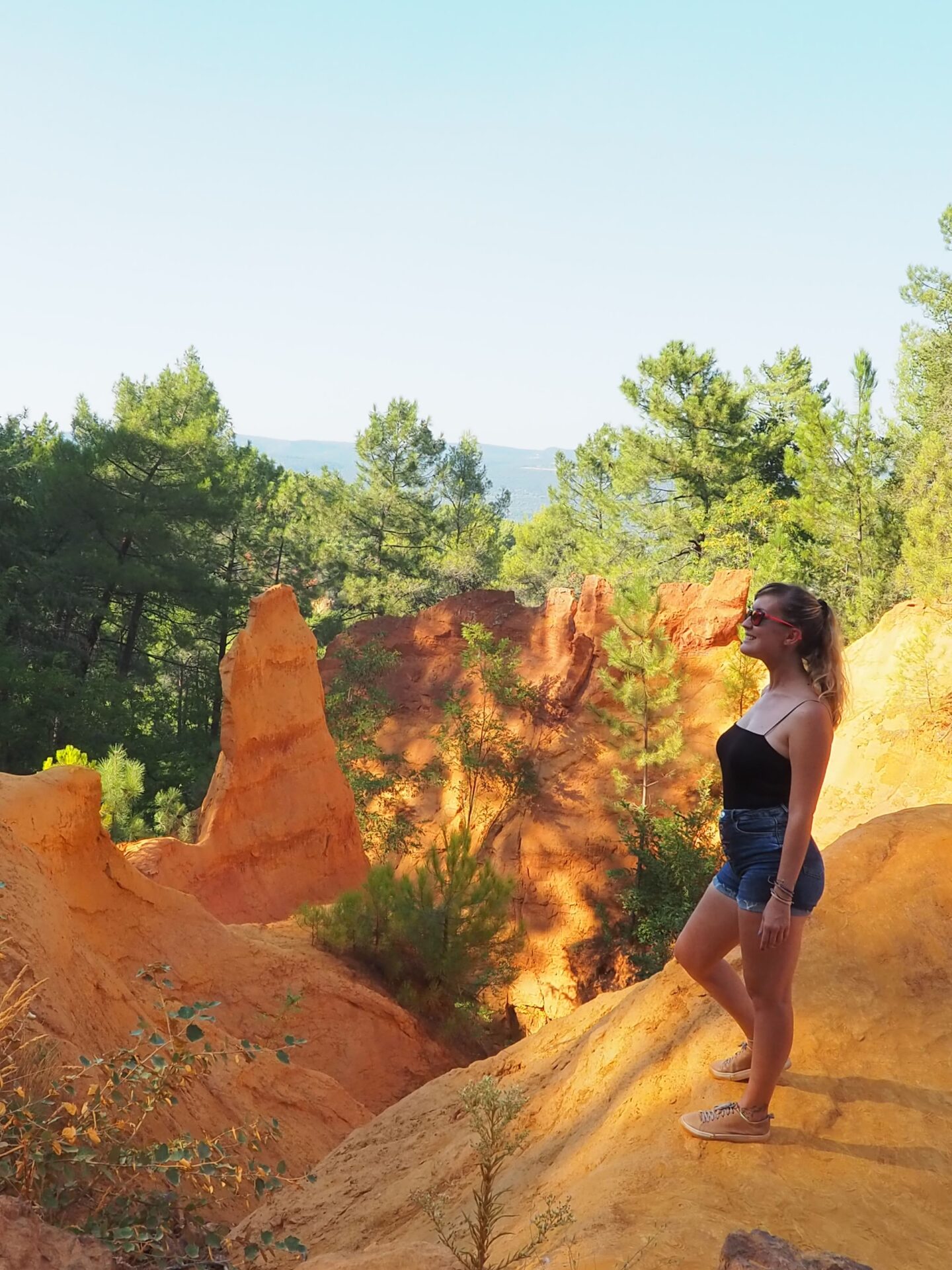 Chica sonriendo en el Sendero de los Ocres (Sentier des Ocres) en Roussillon, rodeada de formaciones rocosas rojizas y pinos verdes en la Provenza.