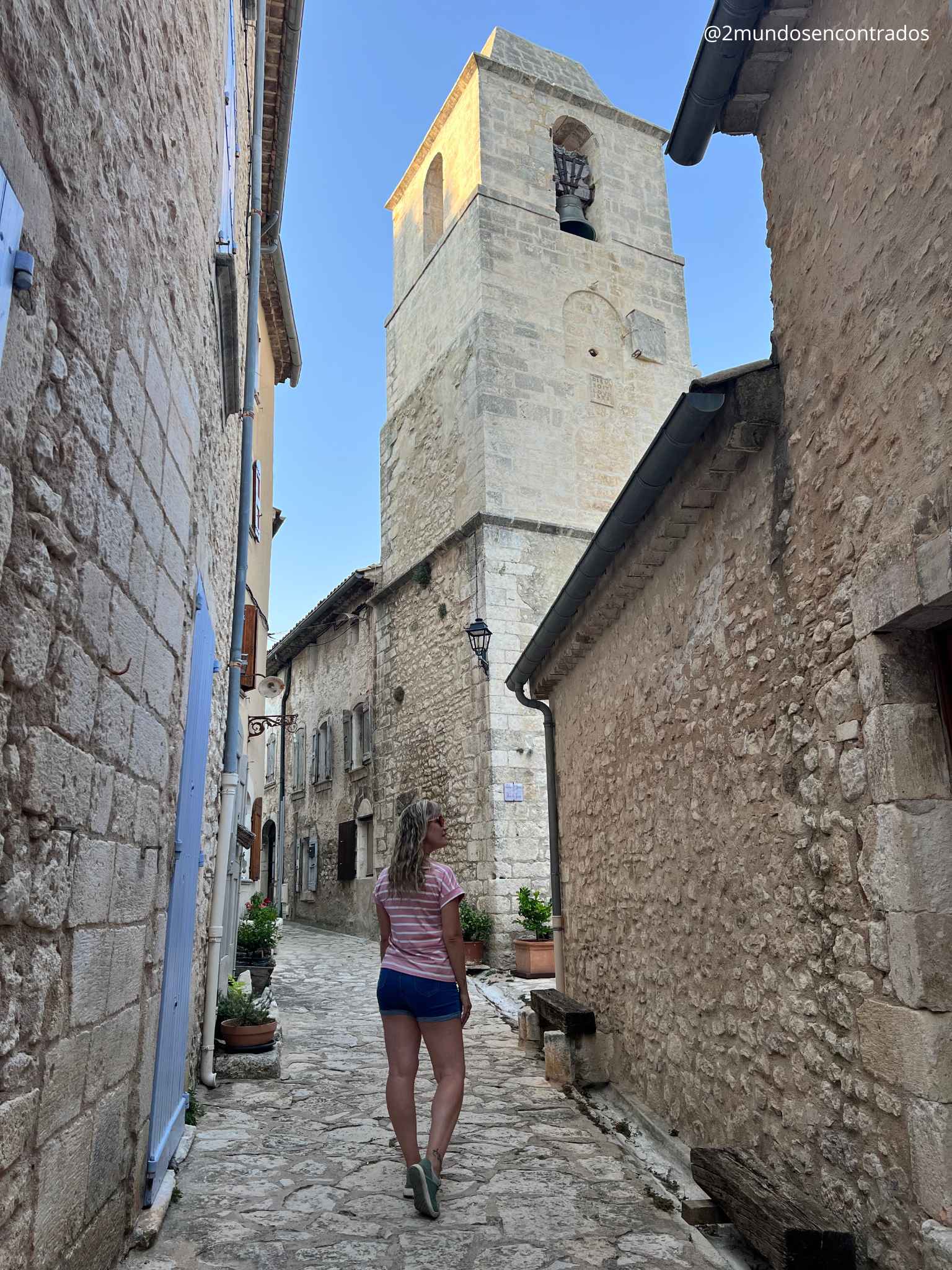 Eva caminando por una calle empedrada del casco histórico de Simiane-la-Rotonde, con el campanario de piedra de la iglesia al fondo, en la Provenza francesa.