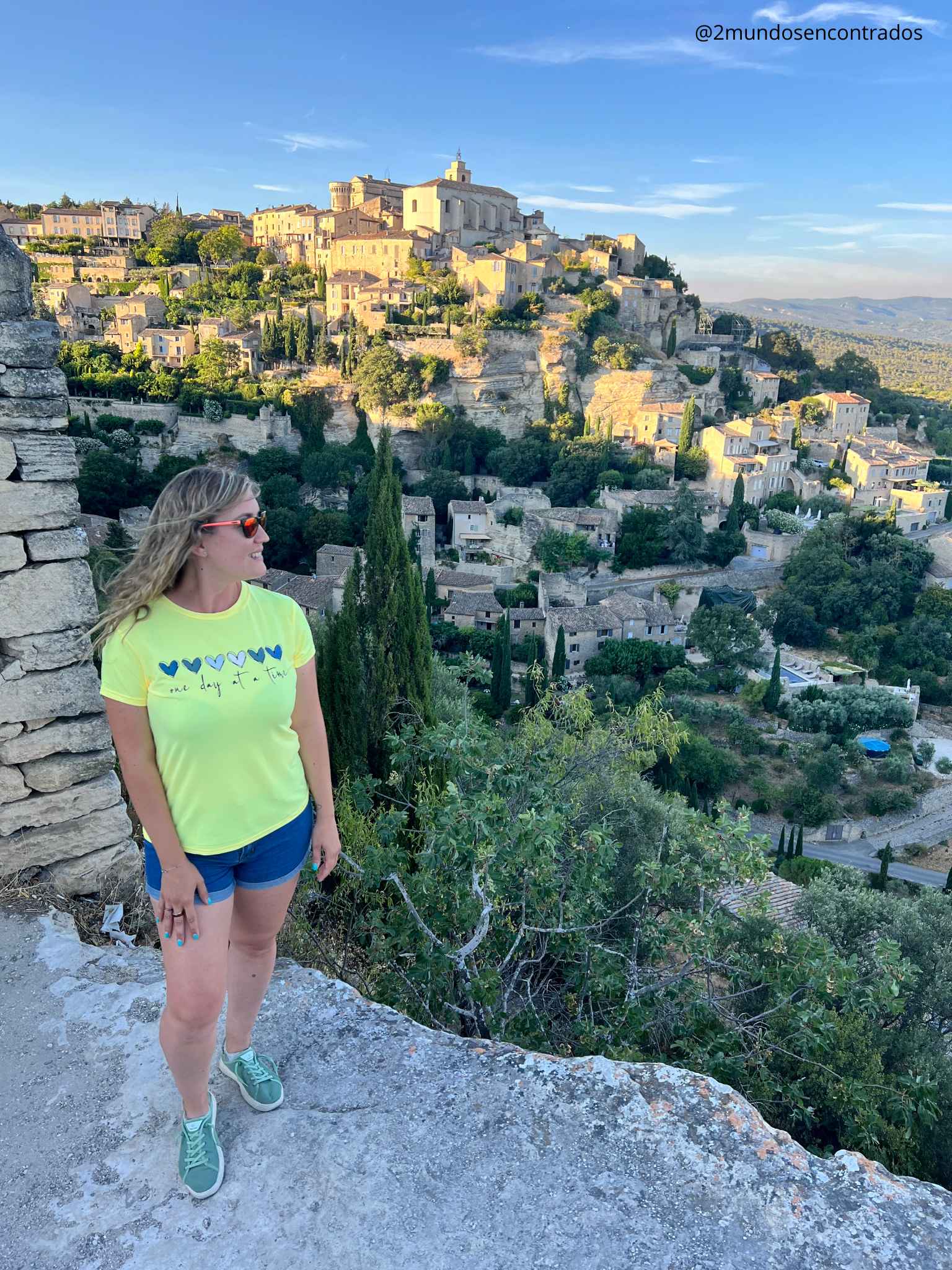 Eva está parada en un mirador de piedra, sonriendo y mirando hacia la izquierda. Al fondo, se extiende el icónico pueblo de Gordes, Francia, colgado en una colina de piedra caliza, con sus casas de color crema y vegetación verde bajo un cielo azul brillante. El sol de la tarde ilumina la escena.