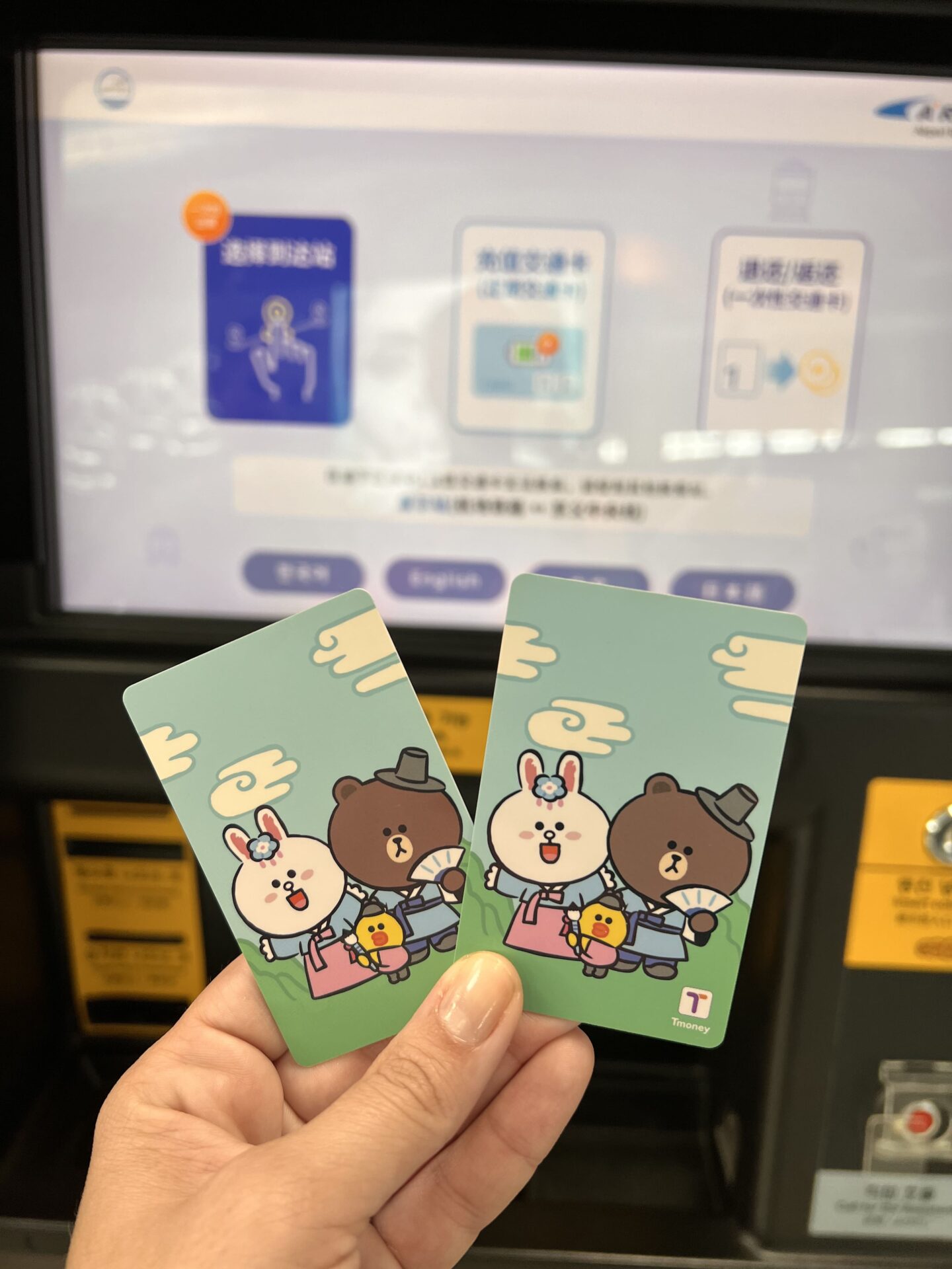 Dos tarjetas T-money con diseño de LINE Friends sostenidas frente a una máquina de recarga del metro de Seúl, Corea del Sur.