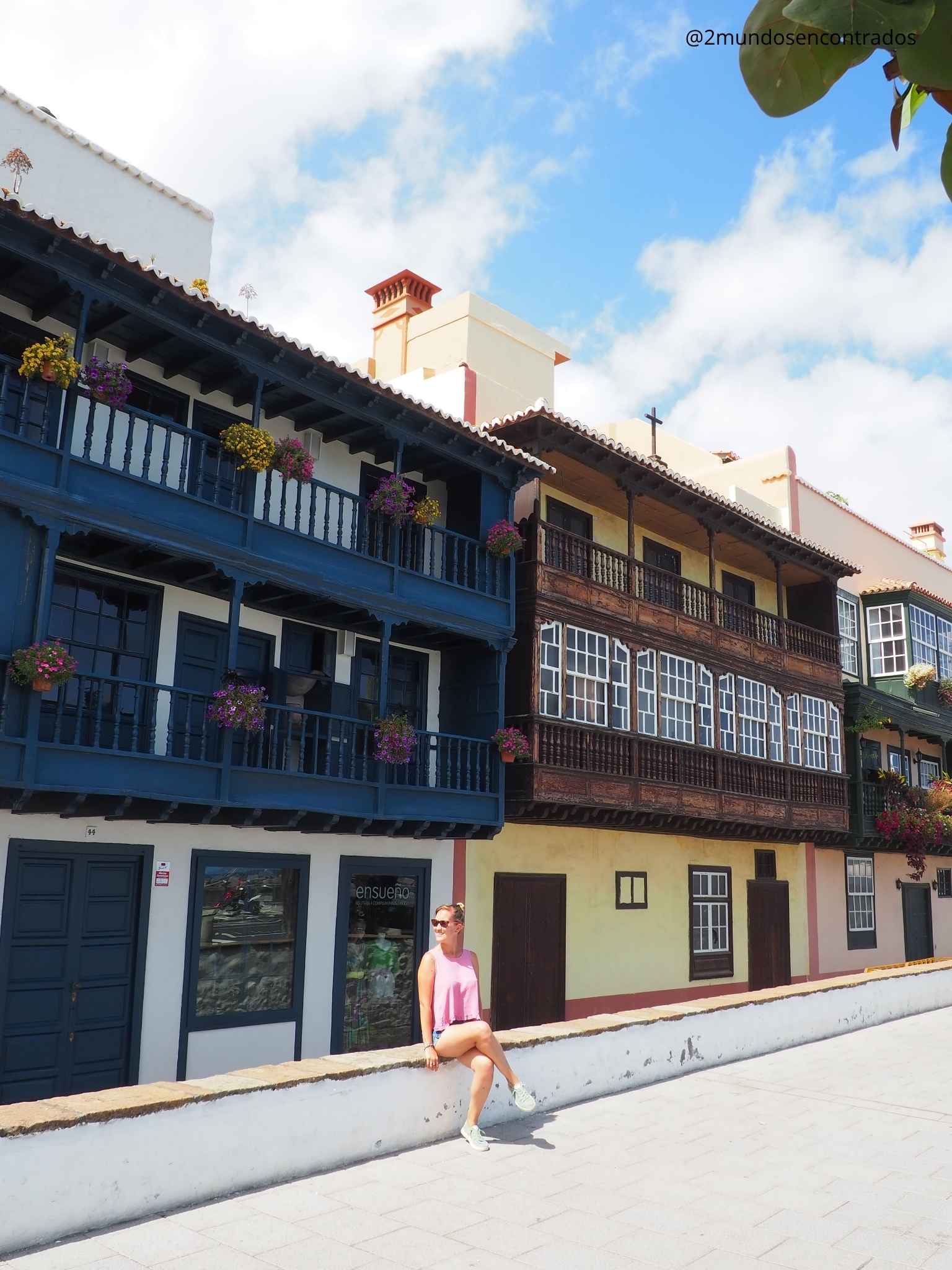 Eva sentada en un muro frente a los tradicionales balcones de madera de colores adornados con flores en Santa Cruz de La Palma.