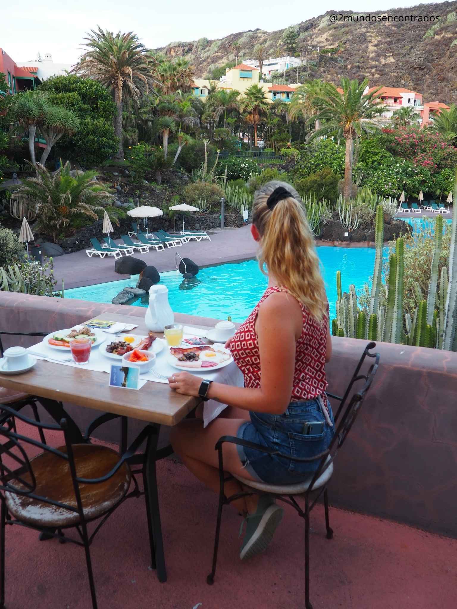 Eva disfrutando de un desayuno al aire libre en la terraza de un hotel con piscina y palmeras en La Palma.